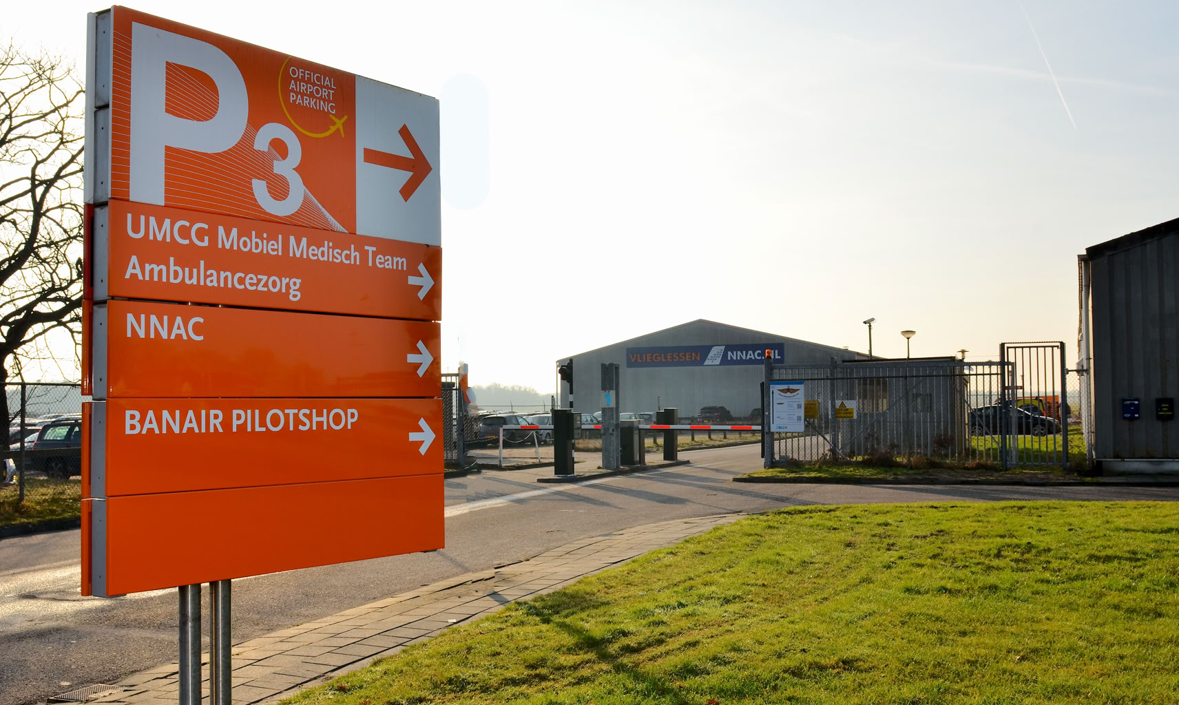 Oranje bord met witte tekst en pijlen, wijzend naar verschillende locaties op Airport Eelde, naast een weg met gras en gebouwen op de achtergrond.